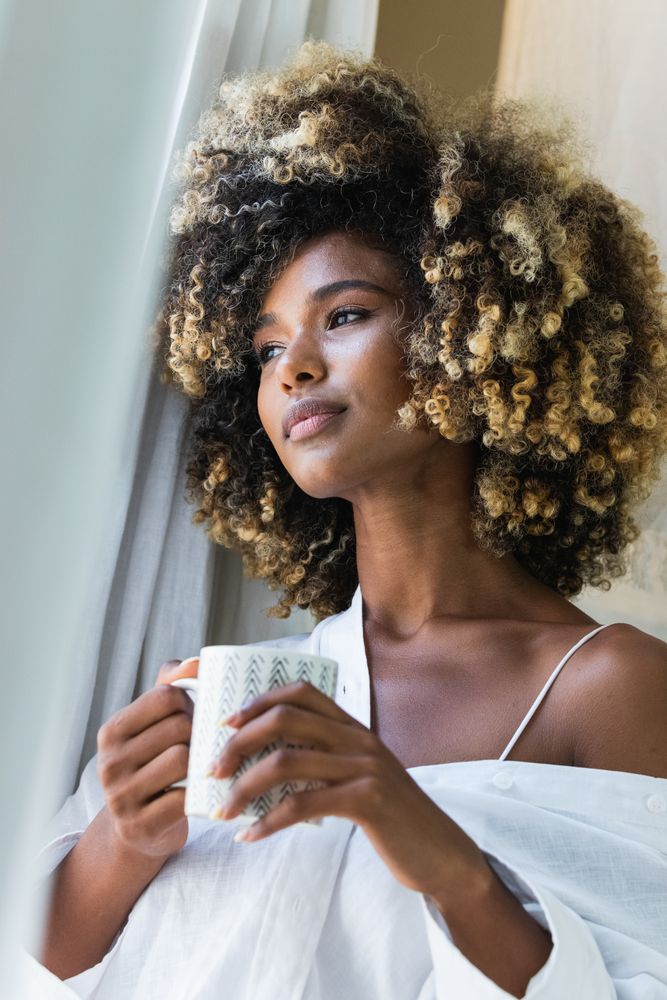 Dreamy Black woman with cup of drink near window