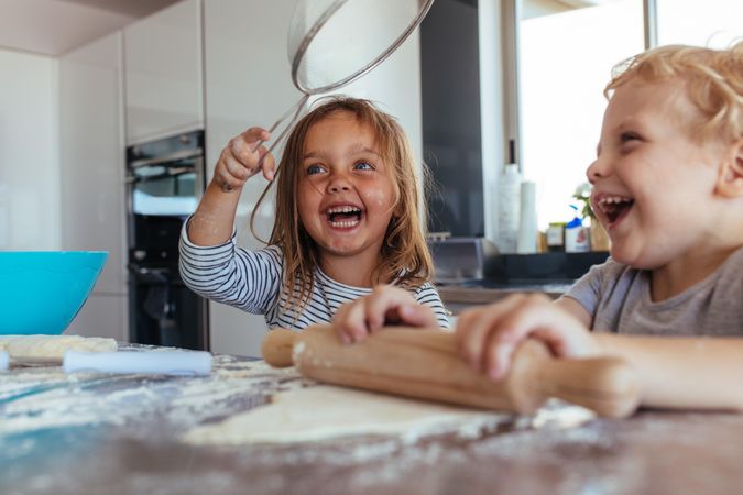 Smiling little girl holding a strainer with boy making dough using rolling pin on kitchen table