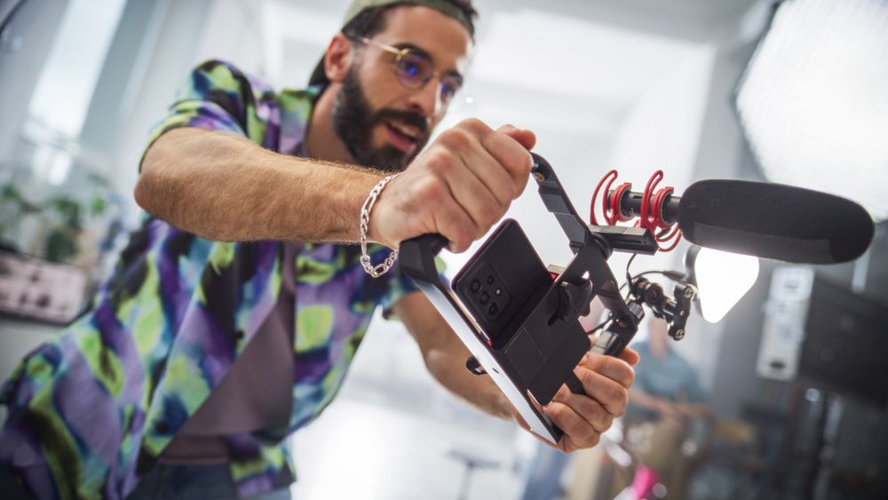 Young Male Filmmaker Operating A Modern Camera With Attached Microphone In A Studio, Capturing Content For A Creative Advertising Project, Showcasing Teamwork And Professional Equipment.