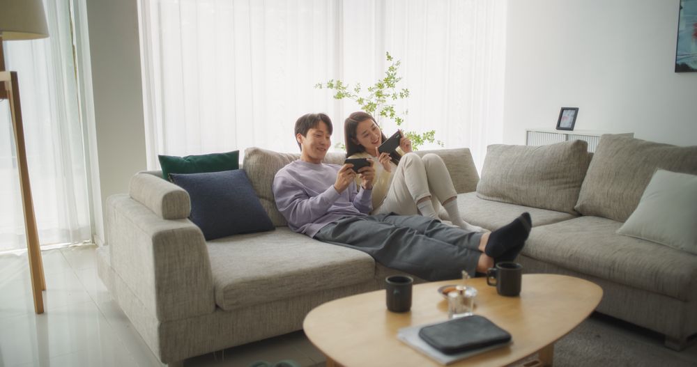 South Korean Couple Sitting on a Cozy Couch, Playing Funny Arcade Video Games Together in a Stylish Bright Living Room.