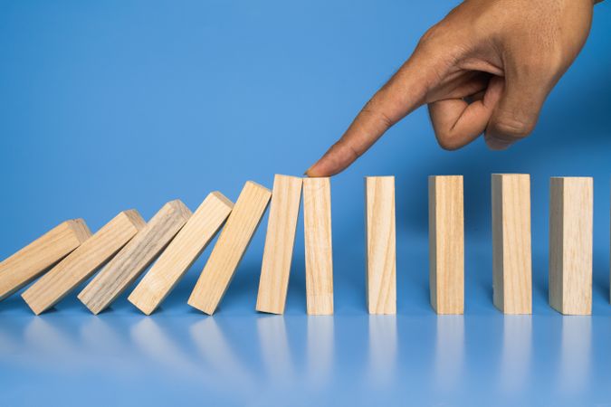 male hand stop falling wooden blocks isolated on blue background. domino effect or risk protection strategy concept