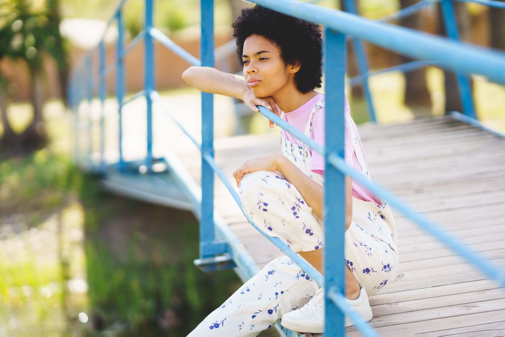 Contemplative woman in floral overalls sitting on bridge over water