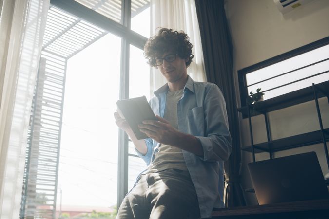 Man working from home perched on desk reading tablet with coffee