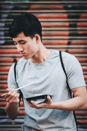 Asian man eating takeaway food with chopsticks on city street