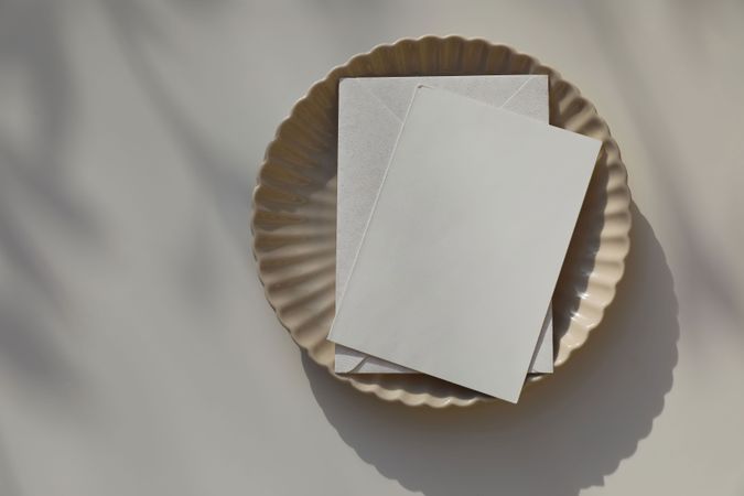 Blank greeting card, invitation mockup, envelope in sunlight. Scalloped ceramic plate on beige table background. Soft shadows. Neutral wedding stationery. Food, restaurant, craft concept, flat lay. Top.