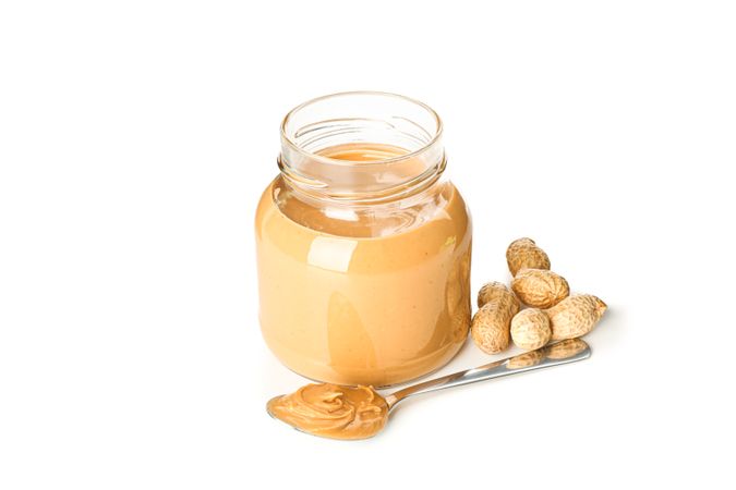 Creamy peanut butter in glass jar, peanut and spoon isolated on light background. A traditional product of American cuisine