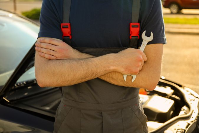 Man in overalls holding a wrench with folded arms. Car inspection