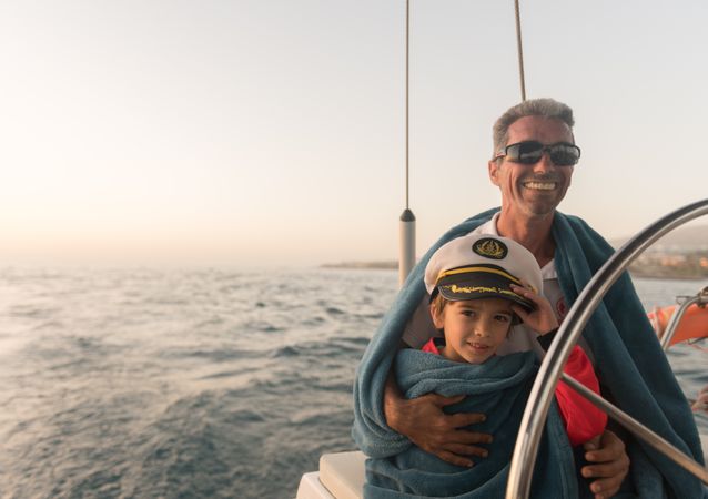 Man hugging smiling child and sitting near steering wheel on yacht in sea
