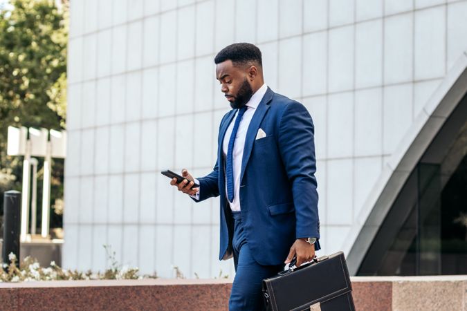 Business Black man in suit leaving the office holding his work briefcase and using smartphone