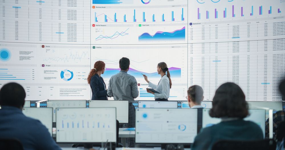 Wide Back Shot of Diverse Team of Data Scientists Discussing Graphs And Reports On Big Digital Screen In Monitoring Office.