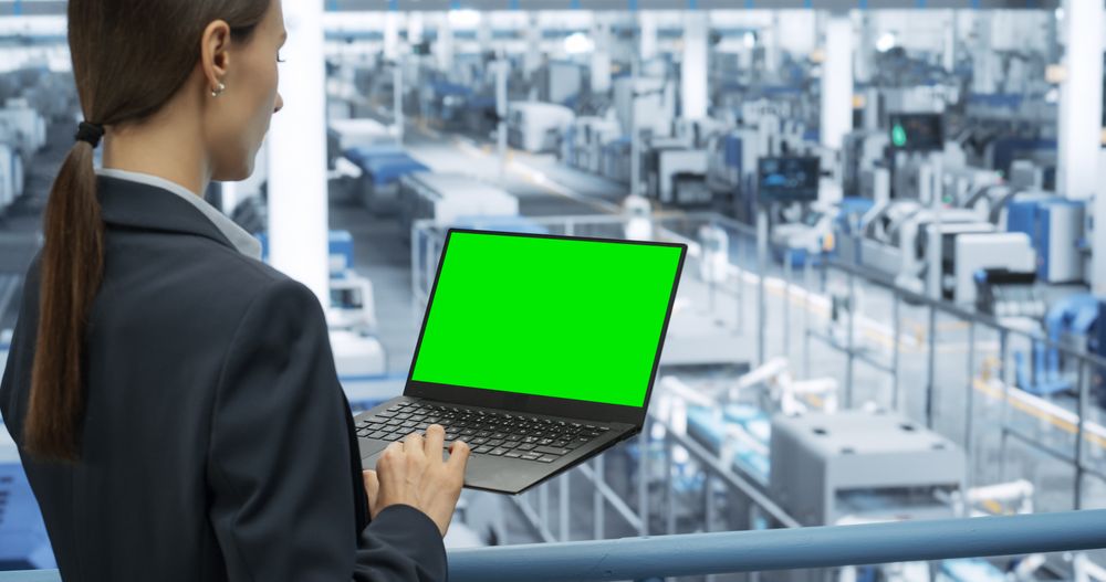 Young Female Engineer Using Laptop Computer with Green Screen Mock Up Display at an Electronics Factory. Automated Manufacturing Machines and AI Robots Are Monitored Online