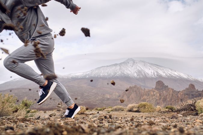 Crop man in activewear running on dirty terrain near mountains