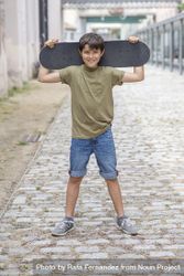 A teenage boy carrying skateboard behind head and smiling 0yX7dL