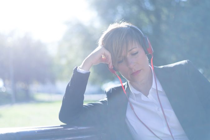 Female in blazer sitting on park bench listening to music on red headphone with sunflare