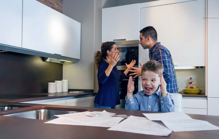 Couple arguing in front of tense son with bills strewn on the kitchen counter