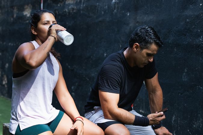Latina woman drinking water near athlete using smartphone