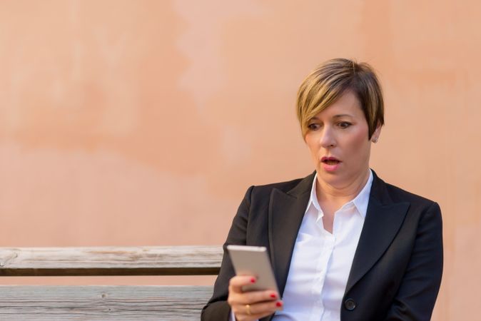 Surprised woman in blazer sitting on bench checking phone in front of peach wall