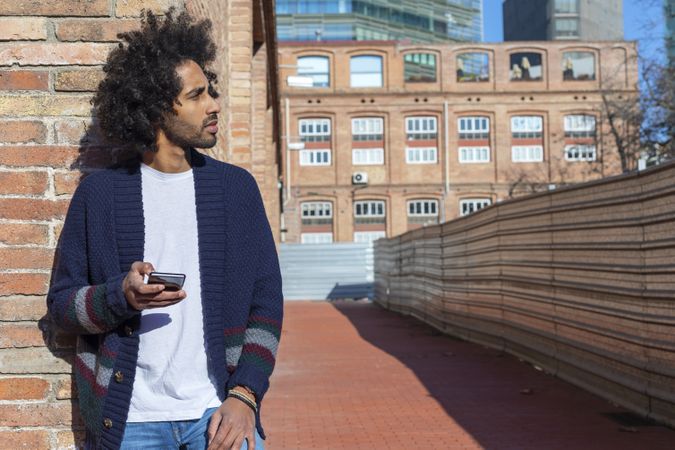 Man using his smartphone with smile while leaning on a brick wall outdoors on sunny day