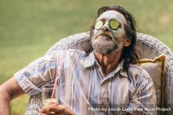 Bearded older man sitting on chair with clay mask and cucumber slice on face 4922Bb
