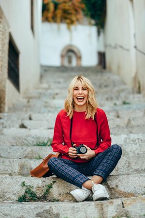 Laughing blonde woman with red shirt sitting on steps outdoors with camera