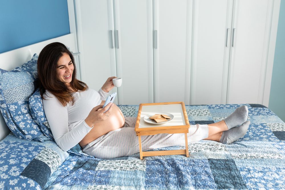 Happy pregnant woman having breakfast and browsing smartphone