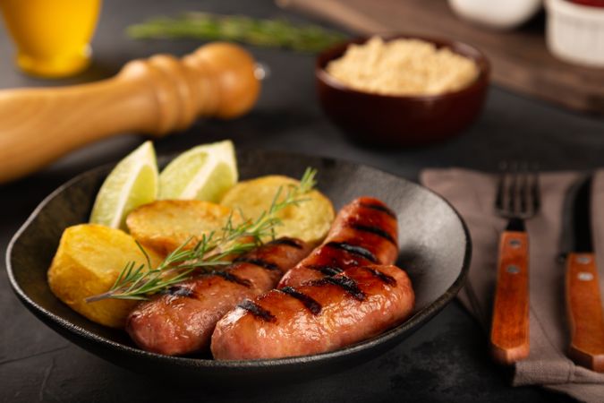 Plate with grilled sausages, potatoes, lime slices and sprig of rosemary on table