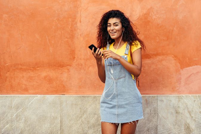 Arab woman with curly hairstyle using phone and headphones in front of on orange wall