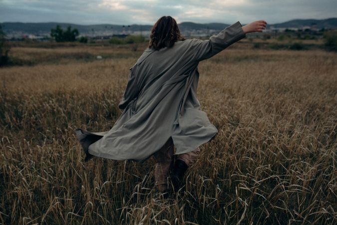 Back view of person in gray coat raising right arm walking on brown grass field