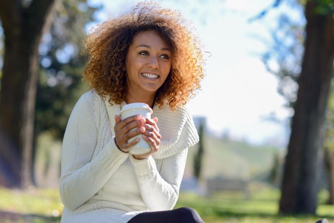 Happy woman looking in the distance while holding warm drink sitting in park