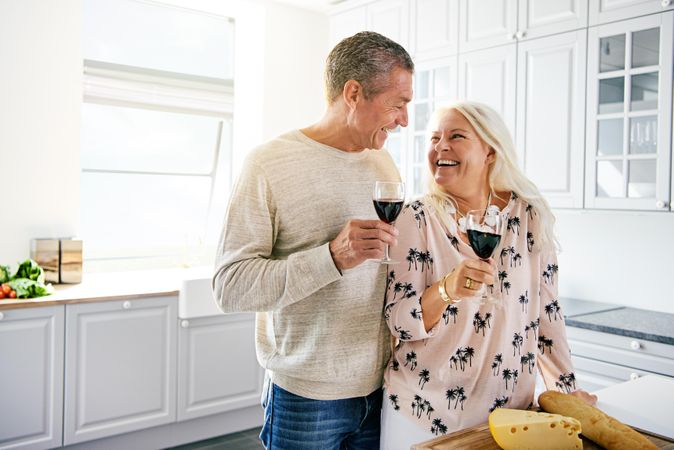 Couple laughing with each other over breadboard with cheese and holding red wine