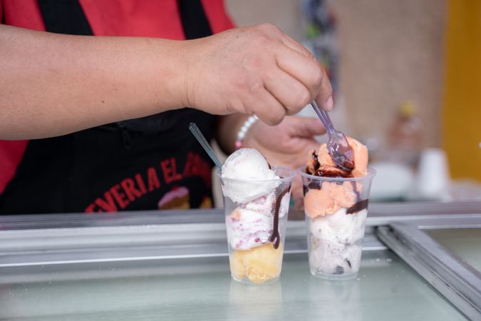 An Hispanic adult woman is serving some ice cream balls with a spoon in plastic cups in the street