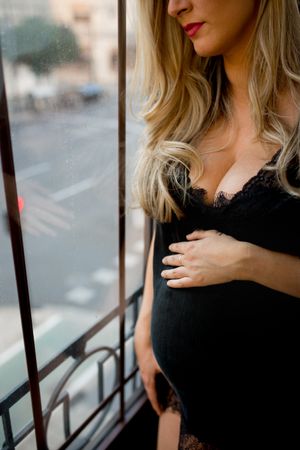 Crop pregnant woman touching belly near window
