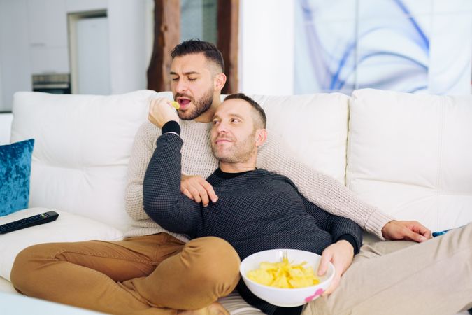 Male couple relaxing on sofa together with bowl of chips