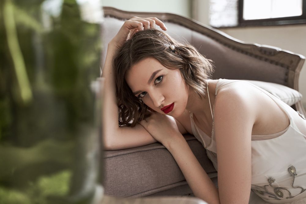 woman with red lips in white dress looking at camera while sitting on floor beside sofa