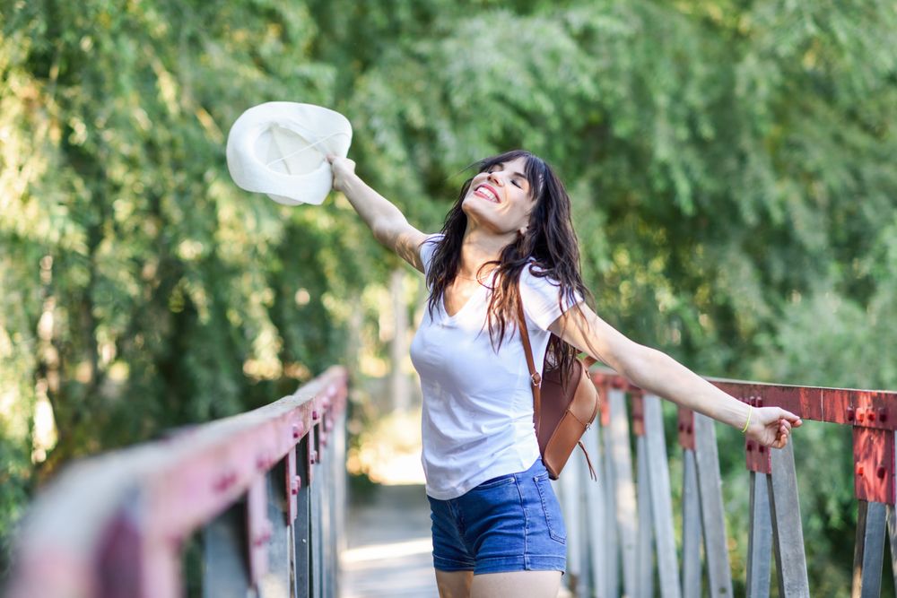 Smiling woman standing on foot bridge in forest with arms opened