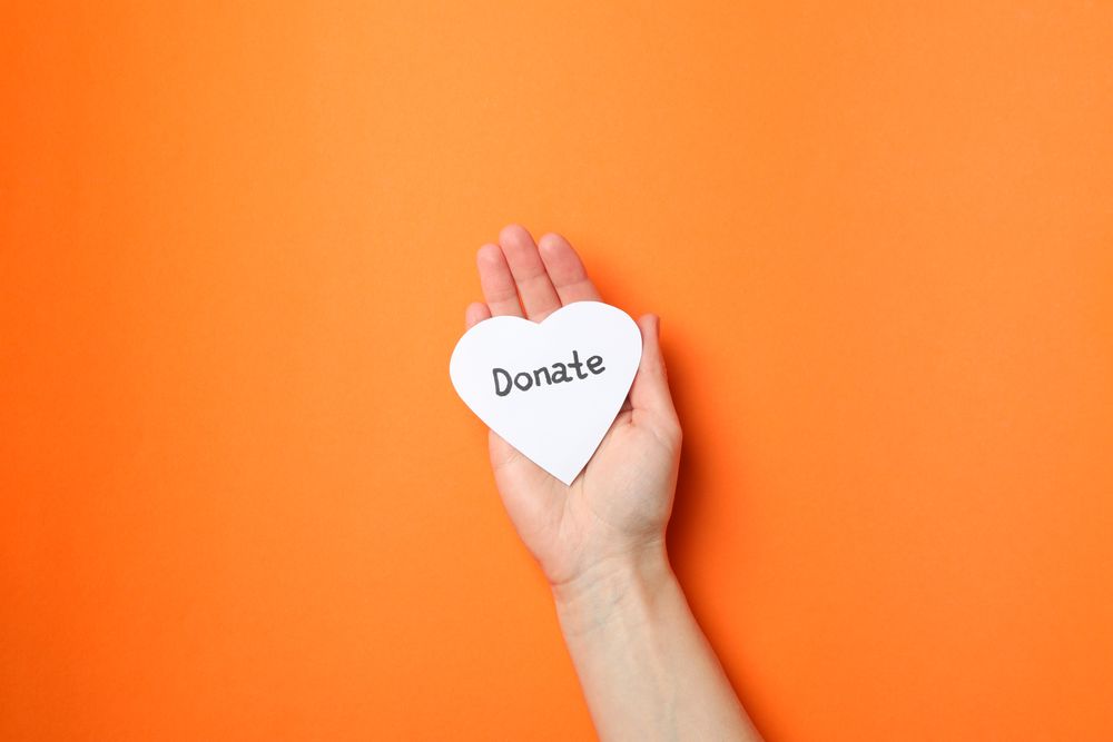 Female hand holds heart with inscription Donate on orange background, top view