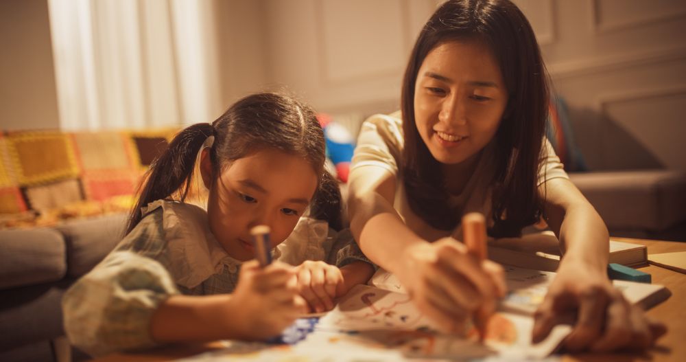 Portrait of Korean Mother Teaching her Little Daughter How to Draw in the Living Room at Home.