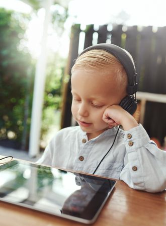 Blond boy using headphones and watching show on digital tablet, vertical