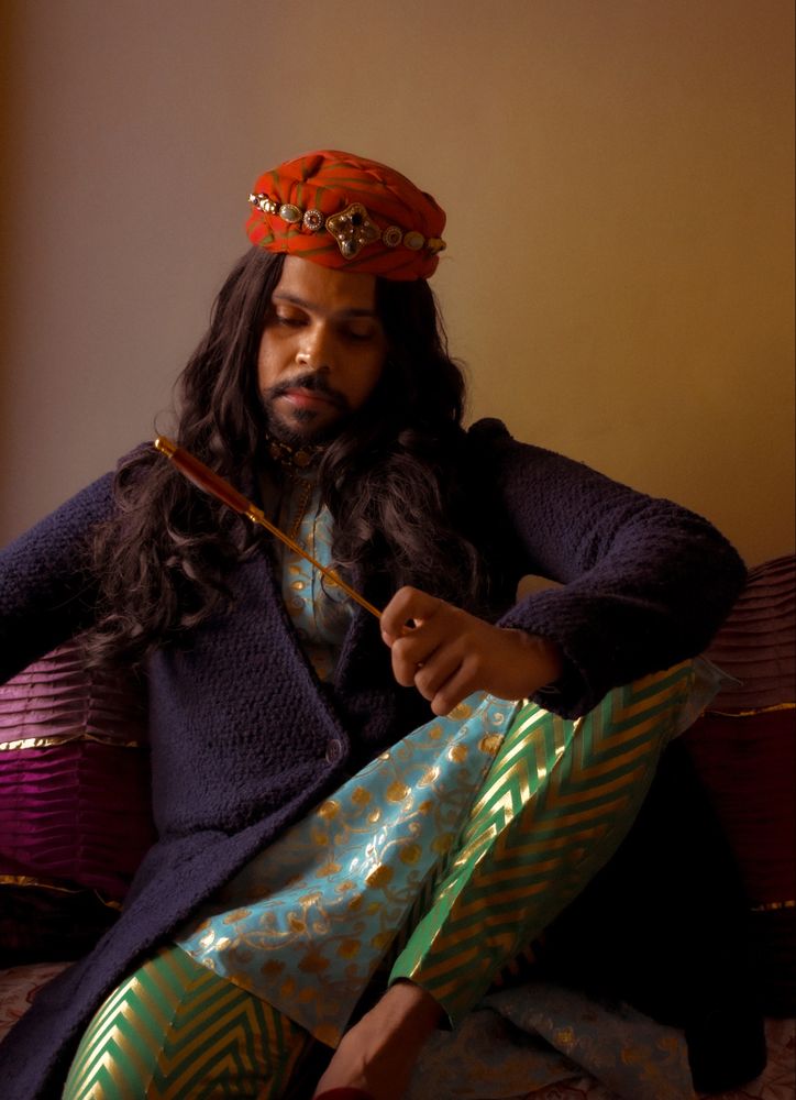Indian man with long hair sitting on floor holding a stick