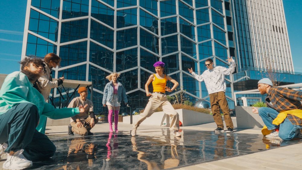 Vibrant Outdoor Scene Of Diverse Young Adults Energetically Dancing In Front Of A Modern Glass Building
