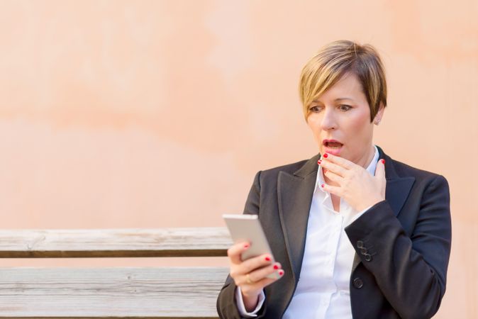 Shocked woman in blazer sitting on bench checking phone in front of peach wall