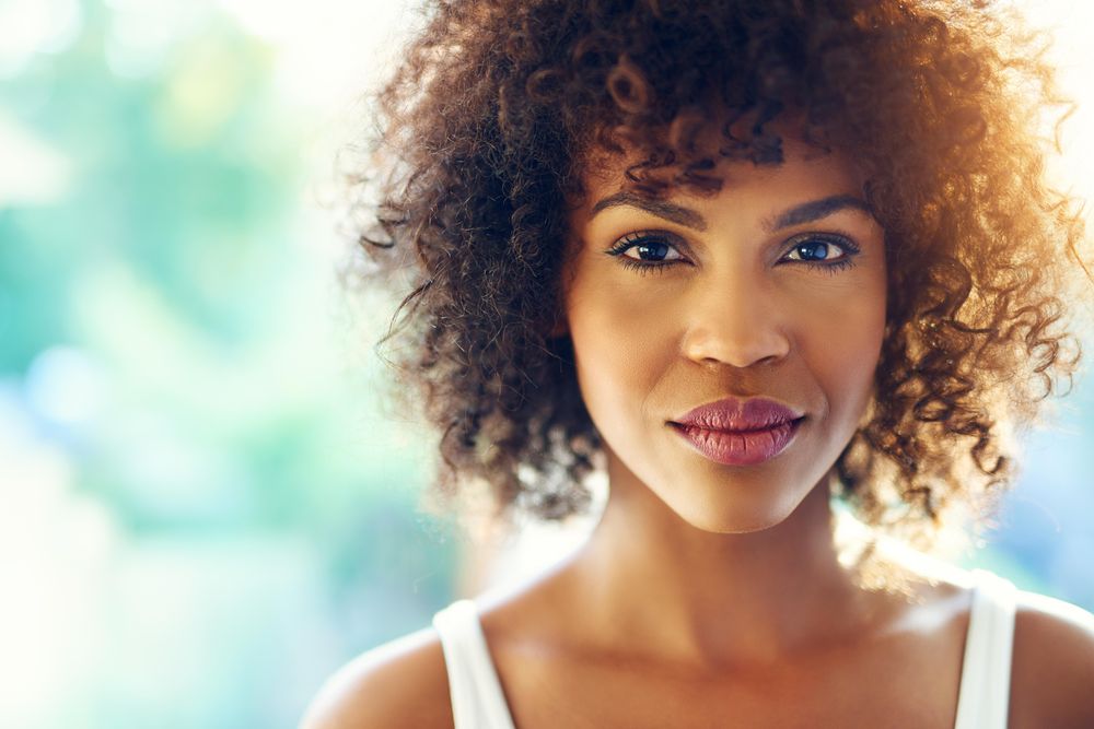 Close up of female with curly hair looking at camera on blurry leafy background, copy space