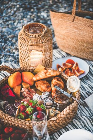 Thatched trey of breads, meats, spreads, and fruit at outdoor picnic with wine glasses