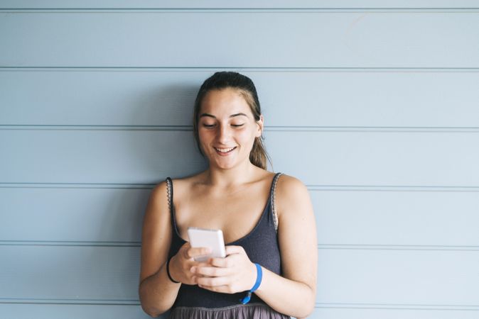 Happy teenage female texting on cell phone while leaning against wood paneled wall