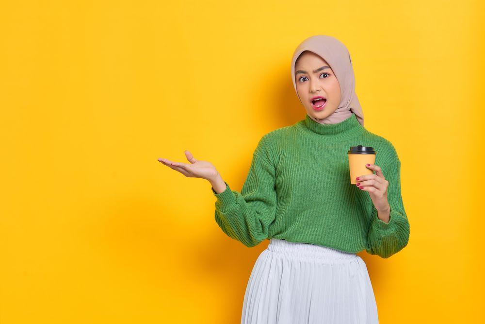 Surprised woman in headscarf holding to go coffee cup and gesturing to the copy space