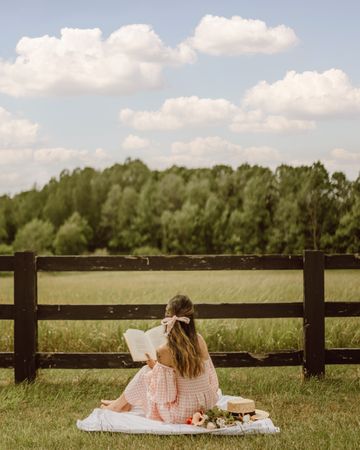Back view of woman in pink dress sitting on grass field reading a book