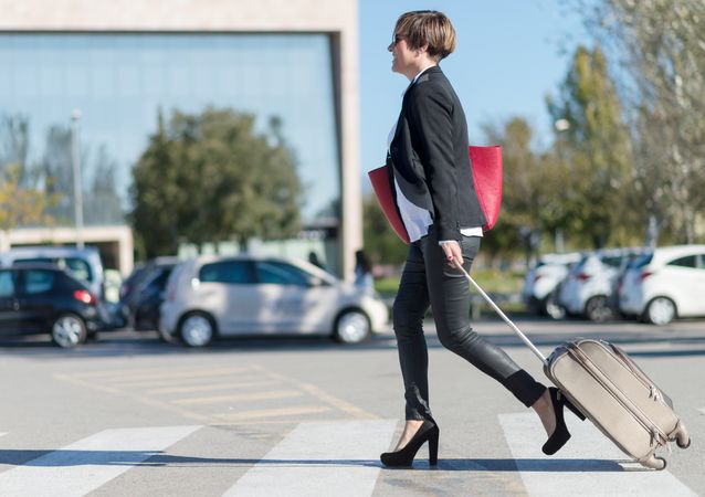 Female in heels walking confidently across street with roller suitcase behind her