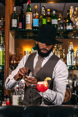 stylish Black bartender in a hat serving a cocktail decorated with garnish. He is scenting the cup with a perfumer