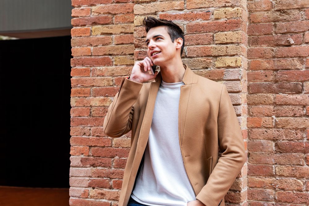 Male in camel coat talking on phone leaning on brick wall