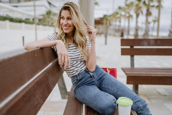 Happy woman with disposable cup of coffee sitting on street bench